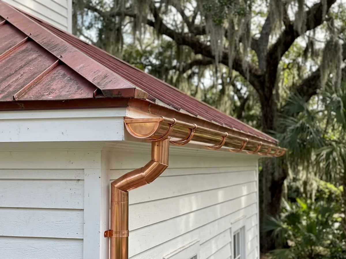 Half-round copper gutter on copper hangers along the eave of a white clapboard house, feeding a round copper downspout